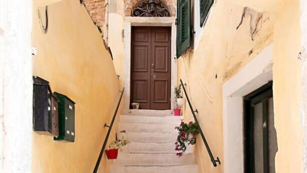 Eine enge Treppe führt zu einer braunen Tür mit grünen Fensterläden, gesäumt von gelben Wänden und Topfpflanzen.