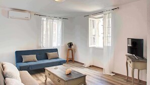 Ein Wohnzimmer mit einem blauen Sofa, einem Holztisch, einem Fernseher auf einem Holzständer und einer kleinen Pflanze auf einem Hocker.