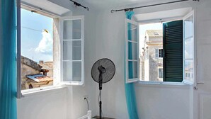 Ein Zimmer mit einem Bett, einem Standventilator und zwei Fenstern mit Vorhängen.