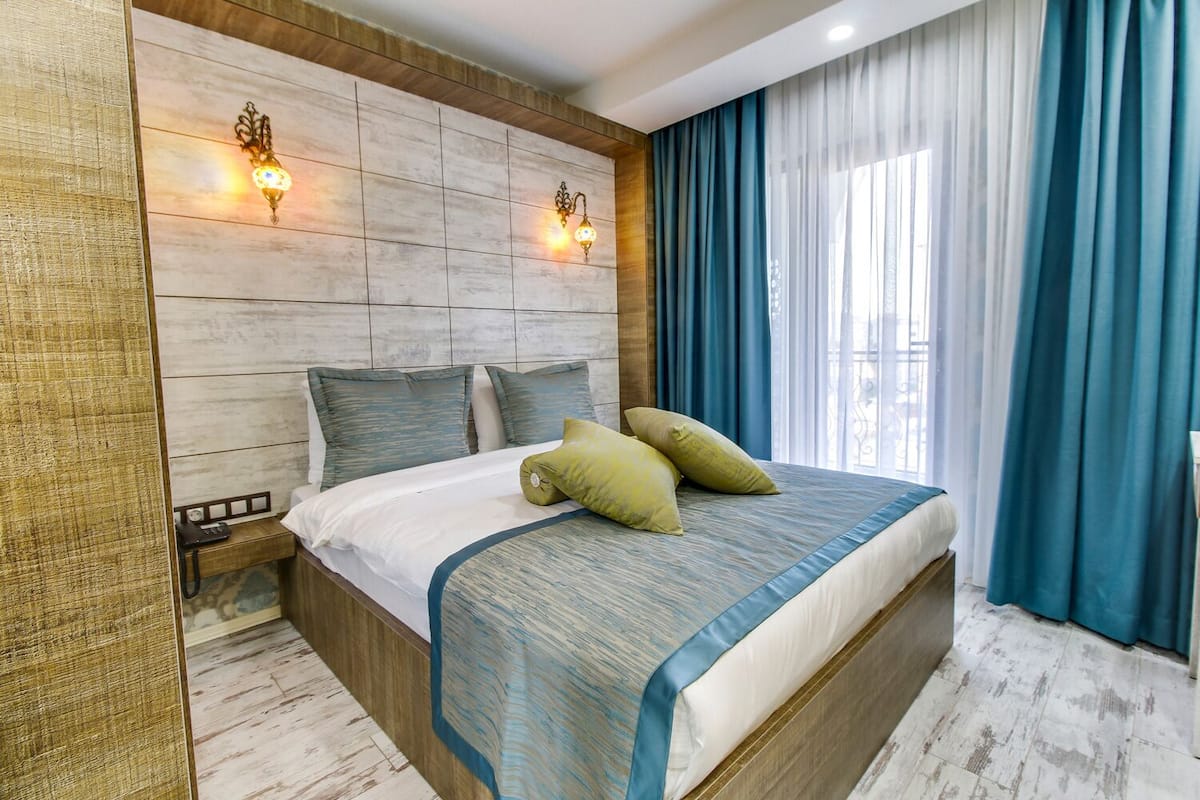 Habitación de hotel moderna con una cama grande, dos lámparas fijadas a la pared y vistas al exterior a través de una ventana con cortinas azules.