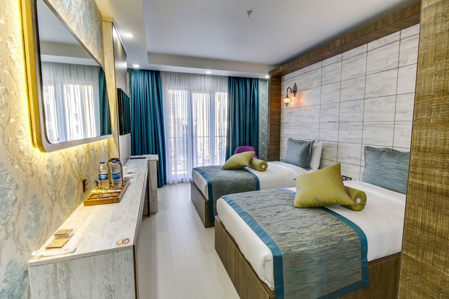A hotel room with two beds, a desk, a mirror, and a wall-mounted light fixture.