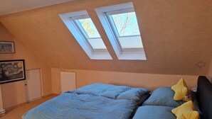 Una camera da letto con soffitto spiovente, un letto con biancheria azzurro e verde, due finestre sul tetto e un quadro appeso alla parete.
