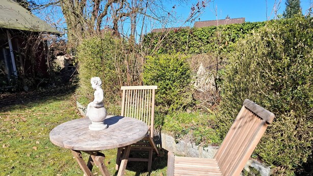 Un tavolo rotondo in legno con una scultura bianca, una sedia di legno e una panchina in un giardino con un siepe e un edificio sullo sfondo.