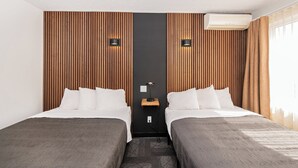 A hotel room with two beds, wooden panel walls, a central nightstand with a lamp, and an air conditioning unit. - Hôtel Althéa Val-D'Or (Val-d'Or)
