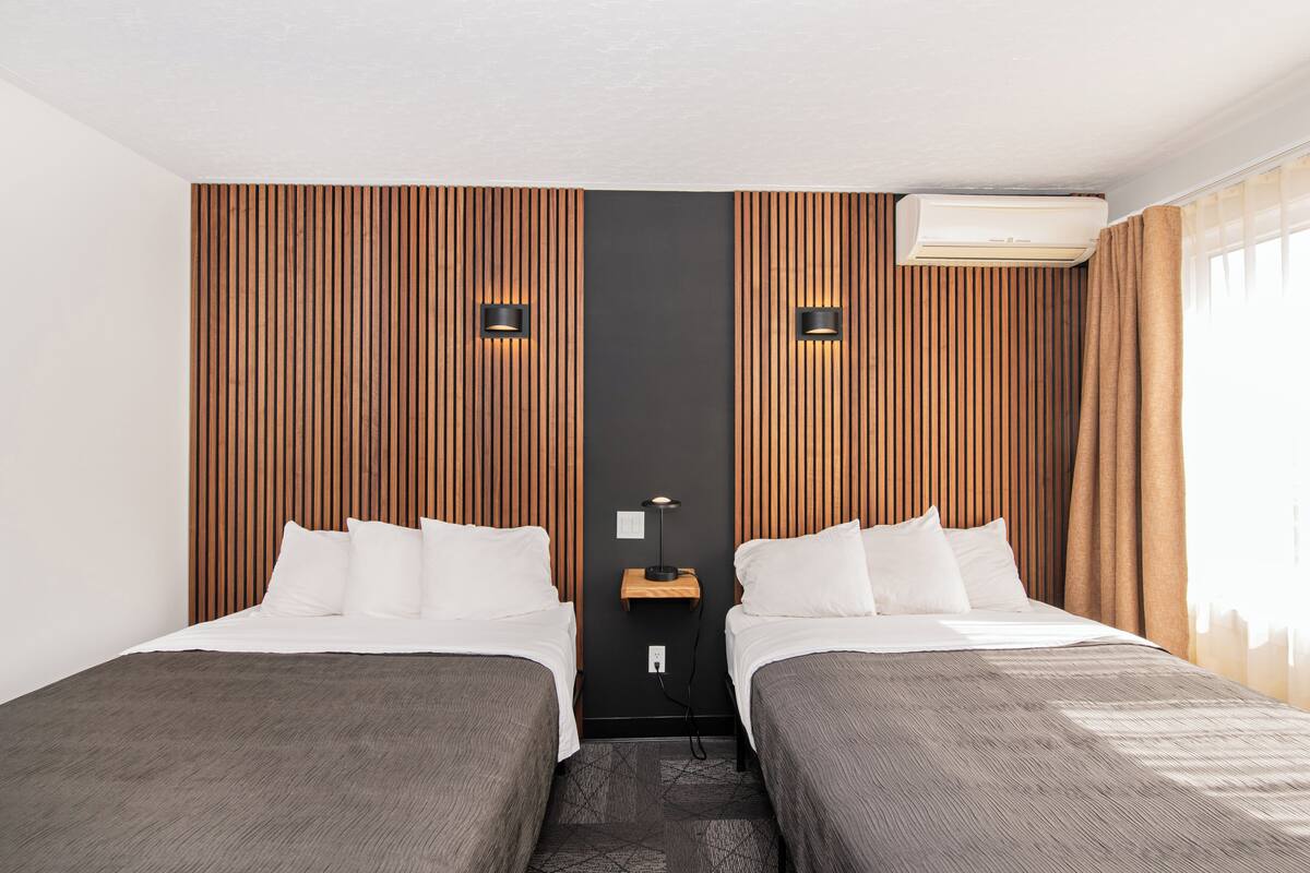 Une chambre d’hôtel avec deux lits, des murs recouverts de lambris en bois, une table de chevet centrale avec une lampe et un appareil de climatisation.