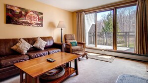 A living room with a brown leather couch, a wooden coffee table, a patterned armchair, and a large window with a view of a wooden deck and trees. - Sunrise - W Glade J-1 (Killington)