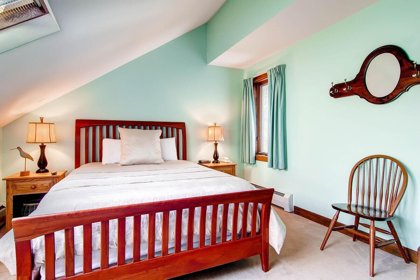 Un dormitorio con una cama de madera, dos mesitas de noche con lámparas, una silla de madera, un espejo y una ventana con cortinas.