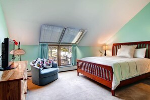 A bedroom with a sloped ceiling, a bed with a wooden headboard, a desk with a computer, a chair with a quilt, and a window with curtains.