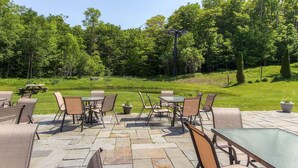 An outdoor patio area with stone flooring, metal tables, and chairs, surrounded by lush greenery and trees. - Sunrise - W Glade J-1 (Killington)