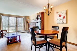 A dining area with a round wooden table, four chairs, a chandelier, and a large window with curtains.