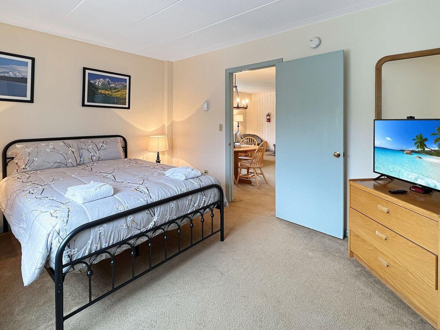 A bedroom with a bed, a television, and a dining area visible through an open door.