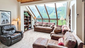 Amplia sala de estar con techo alto, un ventanal con vistas al exterior, un sofá de cuero, un sillón marrón y una estantería.