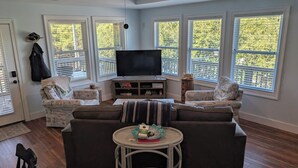 Un salón con un televisor grande, dos sillones estampados, un sofá y una mesita redonda con un control remoto.