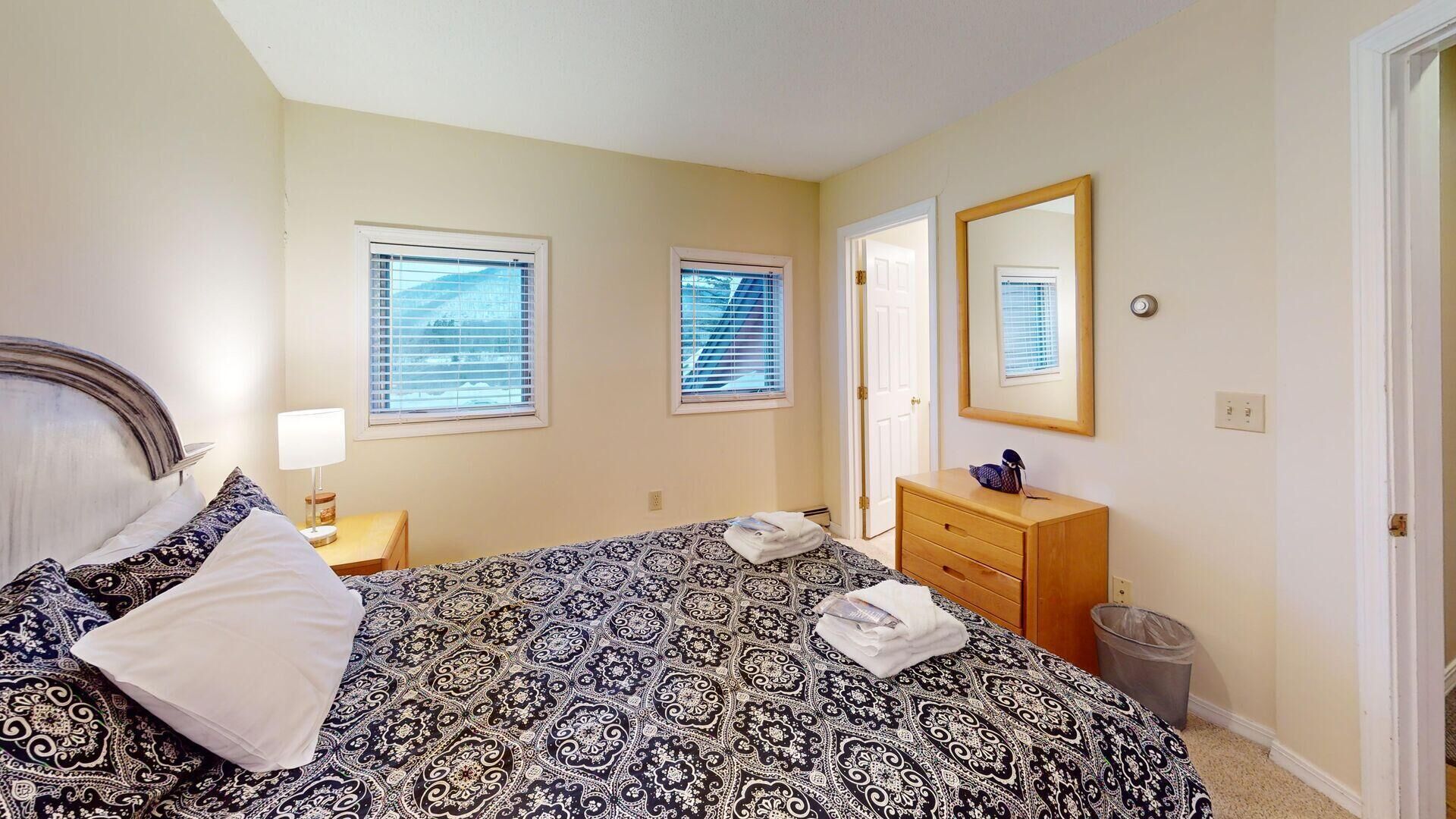 A bedroom with a patterned bedspread, a bedside lamp, a dresser, a mirror, and two windows with blinds.