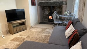 A living room with a stone fireplace, a television on a wooden stand, a grey sofa, and a dining table with chairs. - Tryfan Cottage  -  2.8 miles from Summit of Mt Snowdon (Beddgelert)