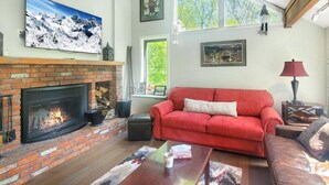 A cozy living room with a brick fireplace, a red sofa, and a large window providing a view of greenery. - Great Escape Ski Lodge (Killington)