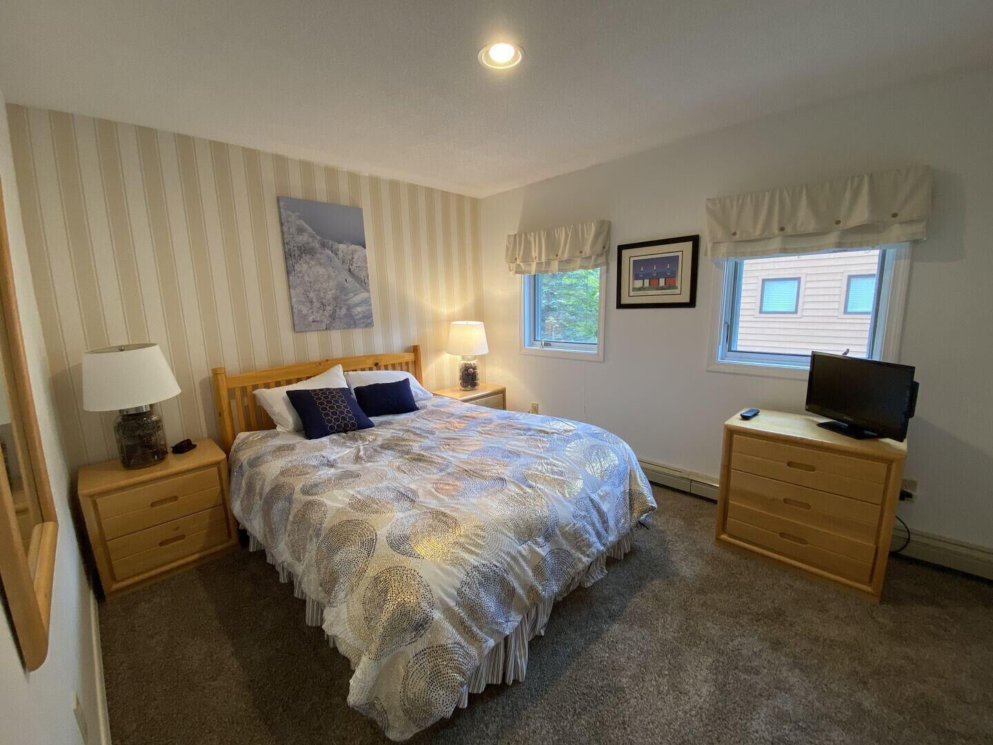 Ein Schlafzimmer mit einem Bett, zwei Nachttischen, einer Lampe, einem Fernseher und einem Fenster mit Vorhängen.