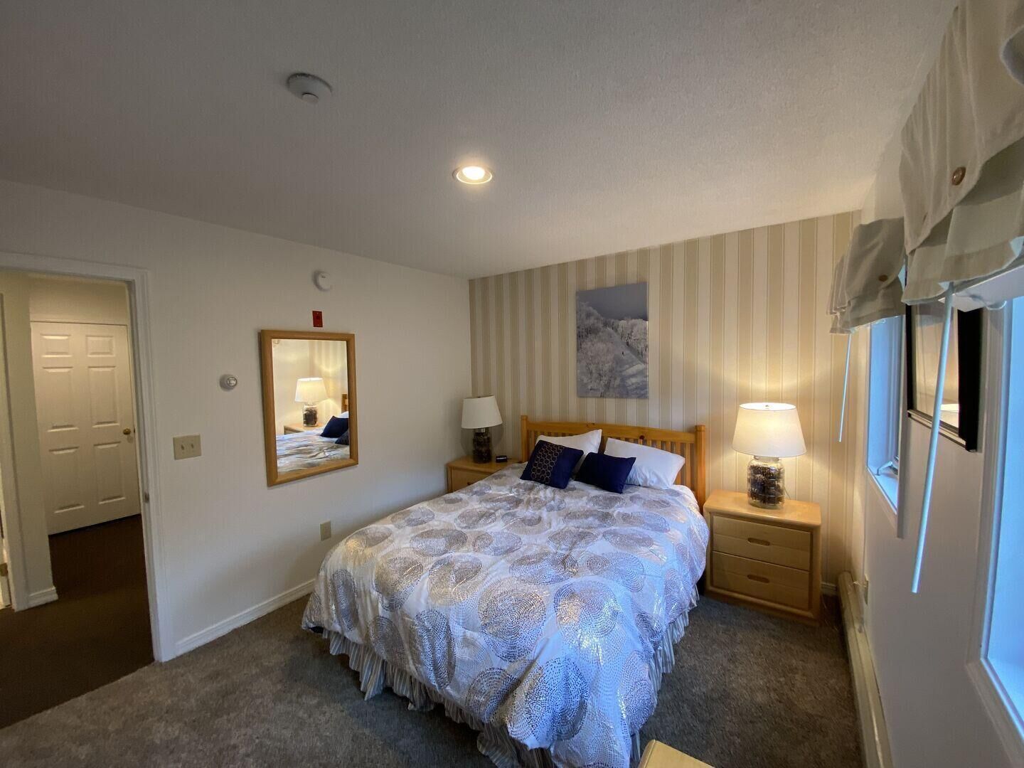 A bedroom with a bed, two bedside tables, a mirror, a window, and a door.