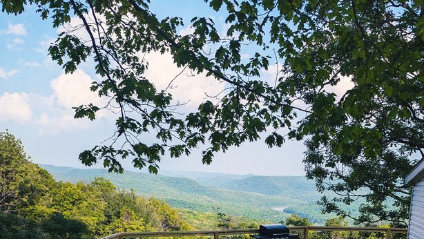 Une terrasse en bois avec un hamac, une table, des chaises et un barbecue, le tout offrant une vue sur un paysage montagneux.