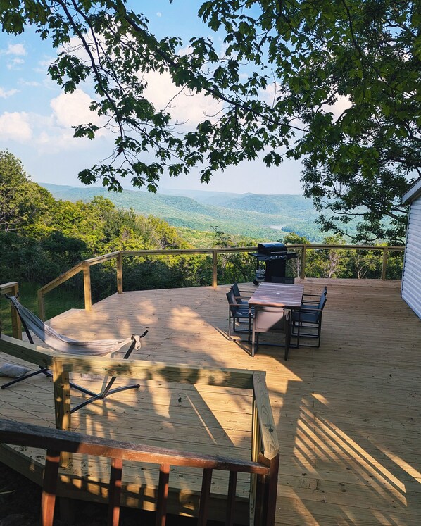 A wooden deck with a hammock, table, chairs, and a barbecue grill overlooking a mountainous landscape.