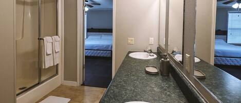 A bathroom with a shower, two sinks, a large mirror, and a bed visible through an open door.