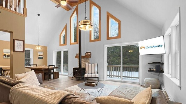 A spacious living area with a fireplace, a dining table, and a large window overlooking a deck. - The Mendon House (Rutland)