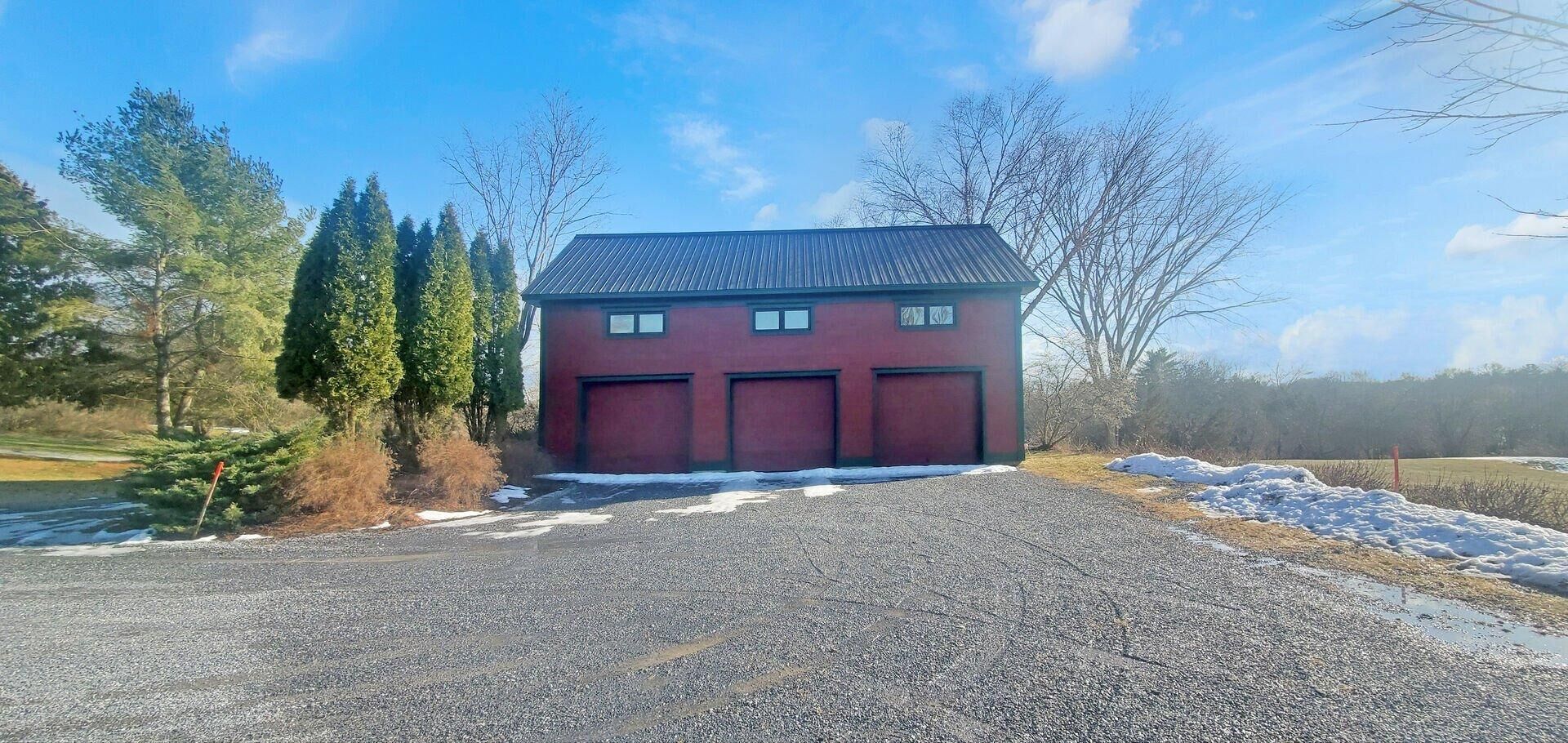 A red building with three garage doors, surrounded by gravel and snow patches, with trees and a clear sky in the background.