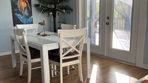A dining area with a white table and chairs, a painting on the wall, a potted plant, and a view through the glass door.