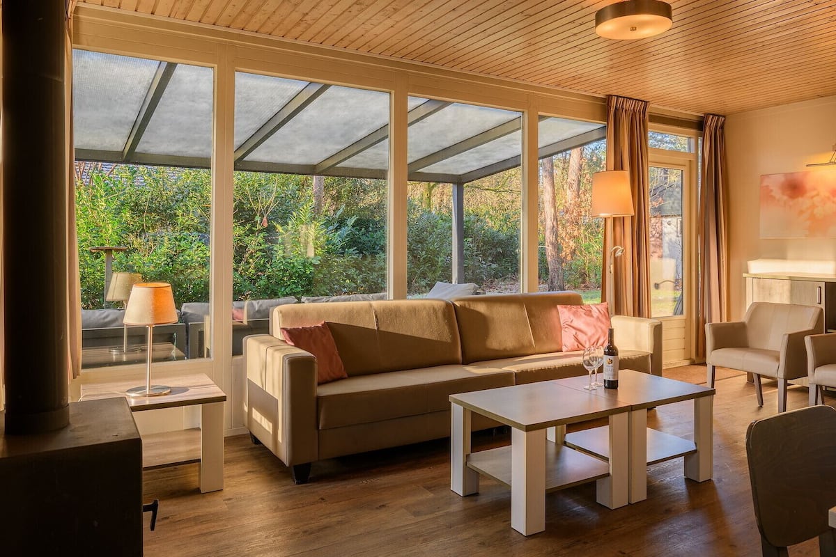 A modern living room with a sofa, coffee table, and chairs. Large windows provide a view of the outdoors.