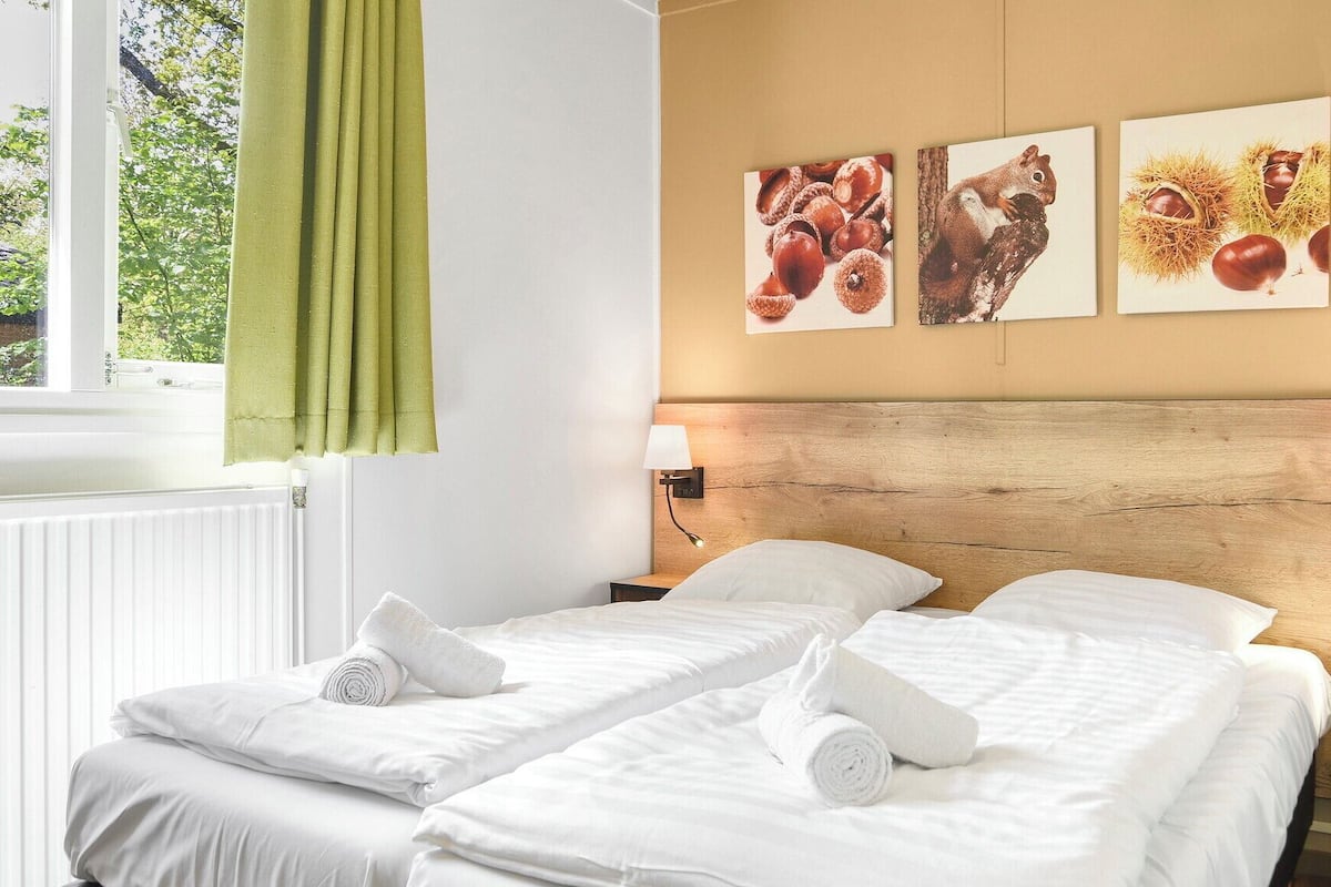 A hotel room with two beds, a wooden headboard, wall-mounted lamps, and three framed pictures depicting nature scenes.