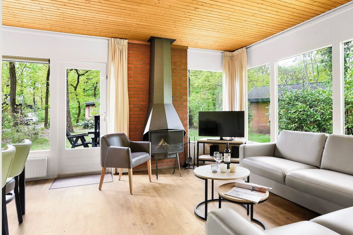 A modern living room with a fireplace, a sofa, a TV, and large windows offering a view of the outdoors.