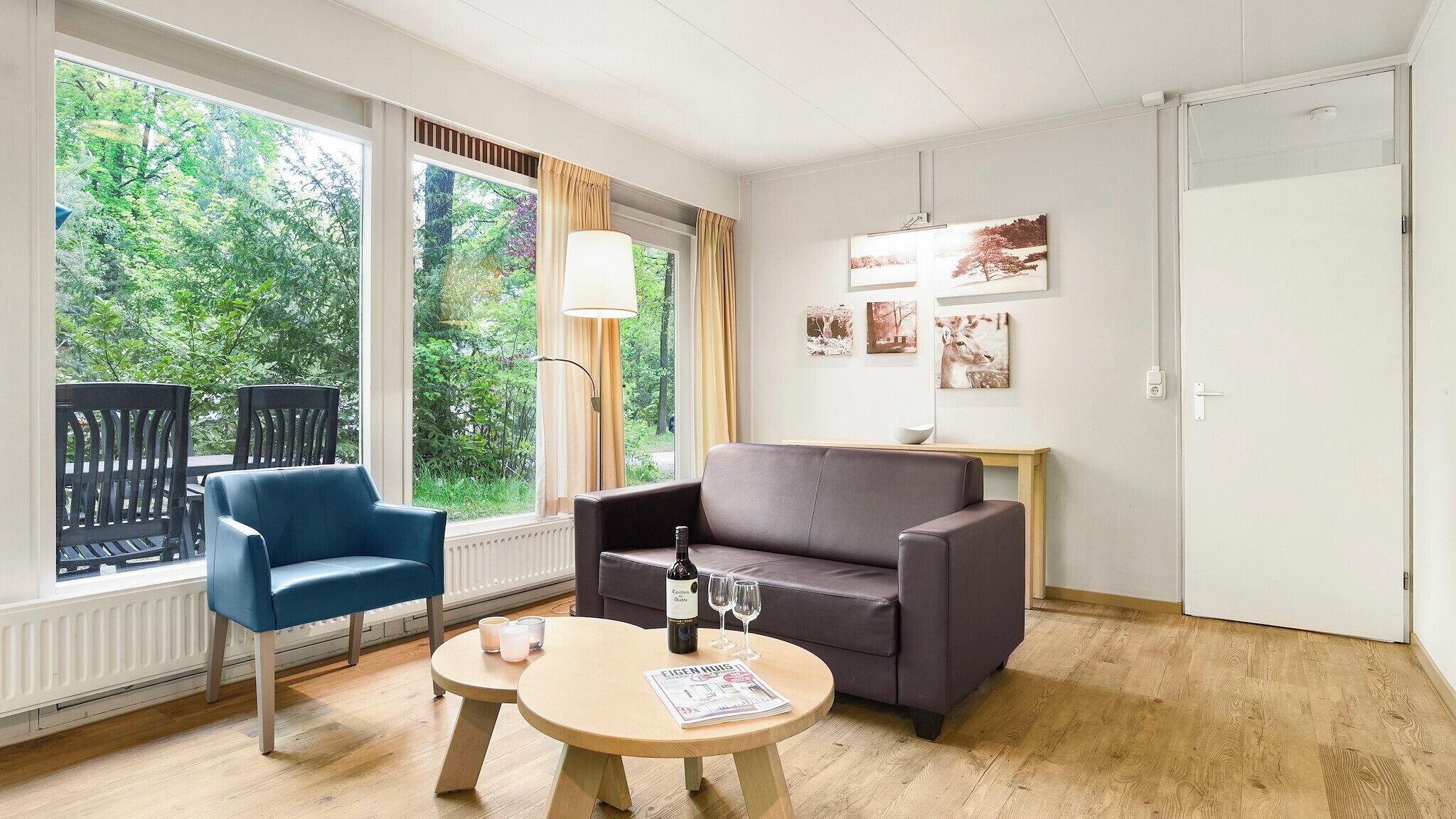 Et moderne værelse med en blå stol, en grå sofa og et rundt bord med en flaske og glas. Store vinduer giver udsigt over grønne områder udenfor.