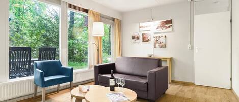A modern room with a blue chair, a grey sofa, and a round table with a bottle and glasses. Large windows offer a view of greenery outside.