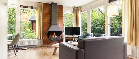 Una sala de estar moderna con chimenea, asientos cómodos, televisor y ventanas amplias con vistas a la vegetación.