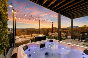 A hot tub with a fire feature, surrounded by outdoor furniture, under a pergola with string lights, overlooking a scenic view.