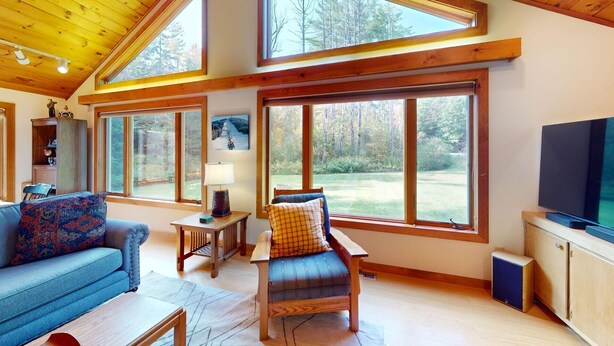 A cozy living room with a sofa, armchair, and a television. Large windows offer views of the outdoors. - The Maple Leaf (Killington)