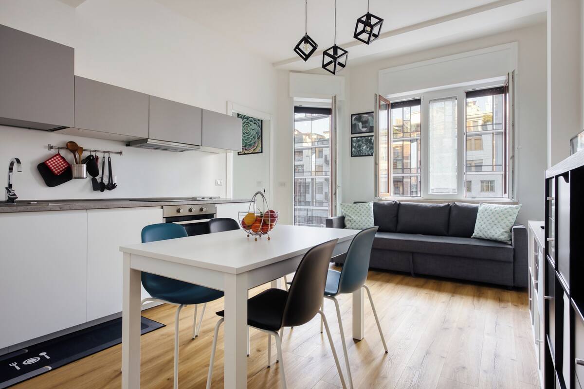 A modern kitchen with white cabinets, a dining table with chairs, and a gray sofa in the living area.