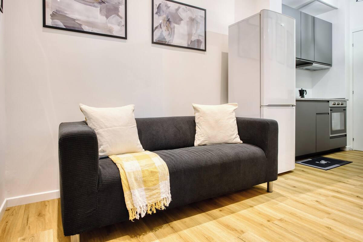 A modern living room with a dark grey sofa, two white cushions, and a yellow and white checkered throw blanket. A kitchen with a white refrigerator, stainless steel oven, and grey cabinets is visible in the background.