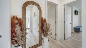 A hallway with a large mirror, white walls, and a wooden floor. There are two vases with dried plants on either side of the mirror. - The Suncatcher by Seaside Vacations & Sales (Chincoteague Island)