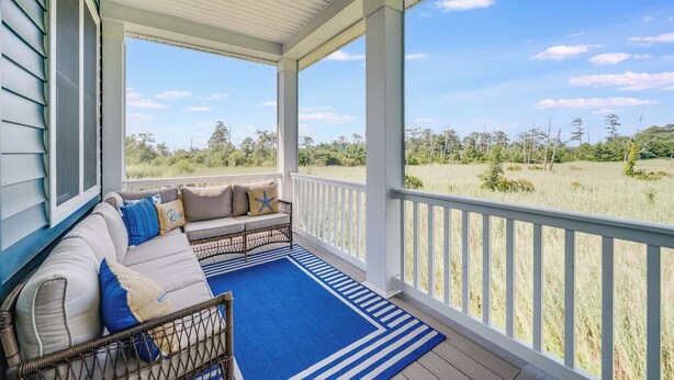 A covered porch with a sofa and a blue rug, overlooking a grassy field. - The Suncatcher by Seaside Vacations & Sales (Chincoteague Island)