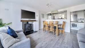 A modern living room with a fireplace, a dining area with wooden chairs, a kitchen with white cabinets, and a flat-screen TV mounted above the fireplace. - Stunning Sea View Blue Haven in Penmaenmawr (Penmaenmawr)
