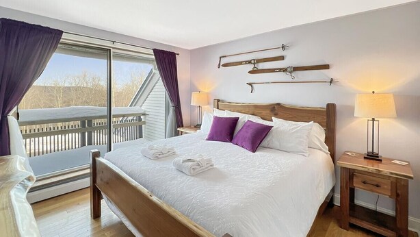 Ein Schlafzimmer mit einem hölzernen Bett, weißer Bettwäsche, violetten Kissen, einem Nachttisch mit einer Lampe und Blick auf einen Balkon.