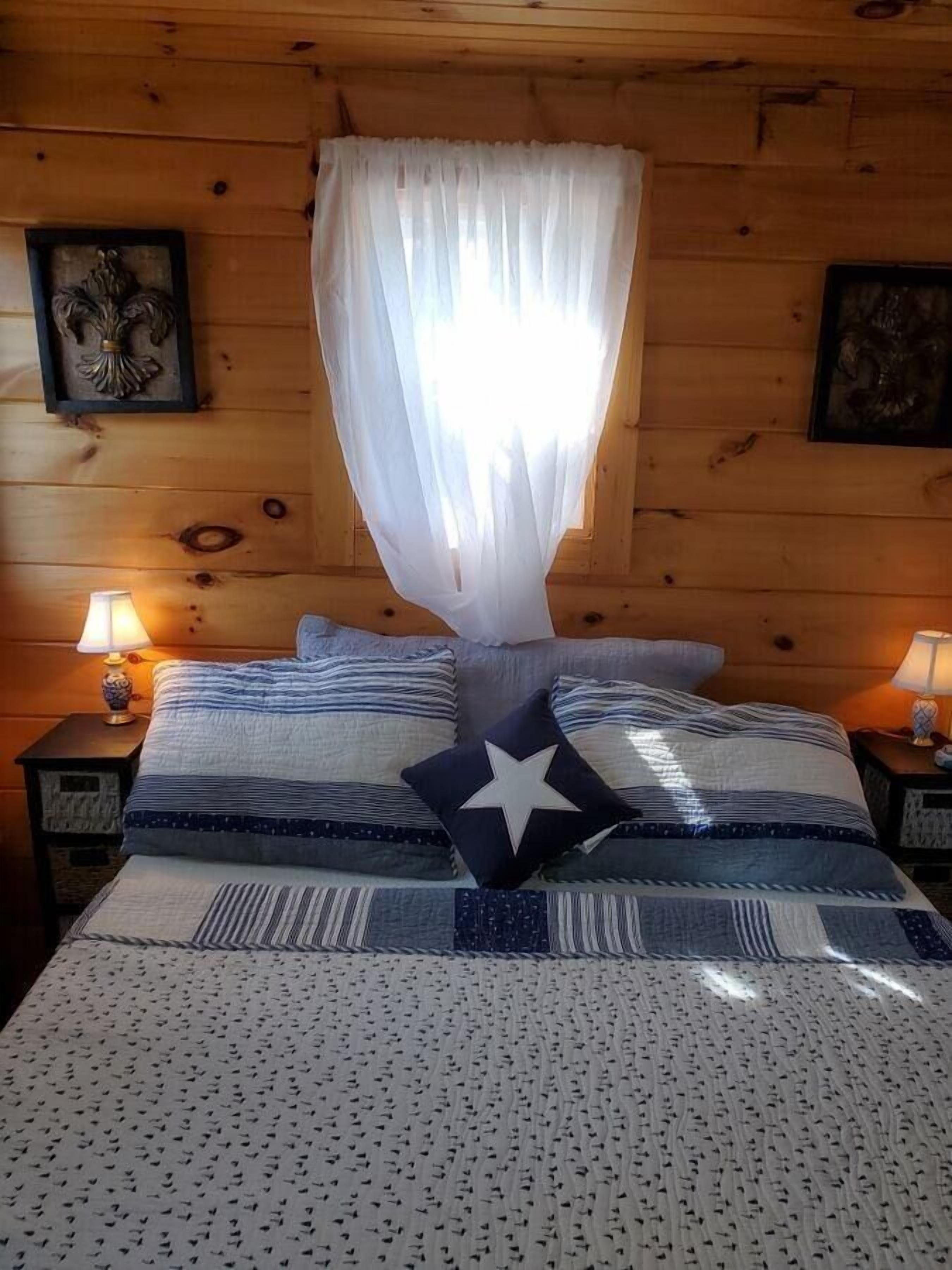 Quarto de cabana de madeira com uma cama, duas mesas de cabeceira com abajures e uma janela com cortinas leves.