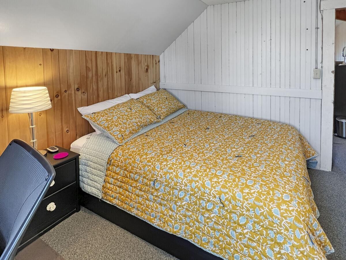 A bedroom with a wooden headboard, a bed with a yellow floral bedspread, a bedside table with a lamp, and a chair.
