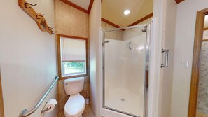 A bathroom with a toilet, a shower, and a window with blinds. - Whispering Pines (Brandon)