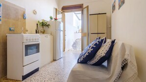 A compact kitchen with a stove, oven, and refrigerator. There is a dining area with a table and chairs, and a sofa with patterned cushions. - Alla Rosa Dei Venti Sud Wind House (Torre Specchia Ruggeri)