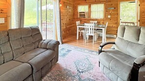 A cozy cabin interior with a sofa, a dining table with chairs, and a window with a 'SURFS UP' sign. - Beach Daze Cottage (York Beach)