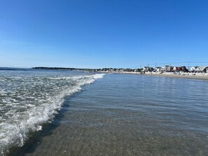 En strand med klart vatten, en sandstrand och byggnader i bakgrunden.