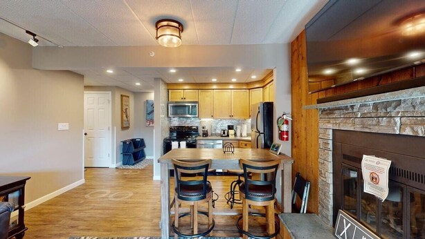 A modern kitchen with wooden cabinets, a central island with bar stools, a stone fireplace, and a large TV mounted above the island. - Mt Green 1-f05 (Killington)