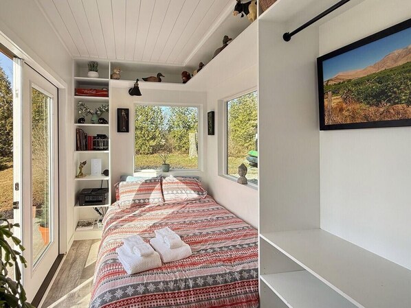 A bedroom with a large bed, a bookshelf, a window with a view, and a mounted picture. - Zen Box Farm (Bomoseen)